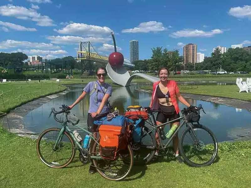 The author and her friend Brenna Forristall ’18 smile while posing with their bicycles in front of a lake with blue sky and fluffy clouds behind them